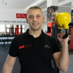 Uśmiechnięty mężczyzna w czarnej koszulce polo z logo siłowni trzyma w dłoni żółty kettlebell na tle sali treningowej.