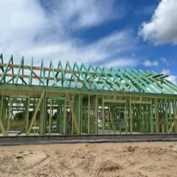 Budowa domu jednorodzinnego w stanie surowym otwartym, widoczna drewniana konstrukcja szkieletowa ścian i dachu, fundament betonowy, plac budowy z piaskiem, w tle błękitne niebo z chmurami.