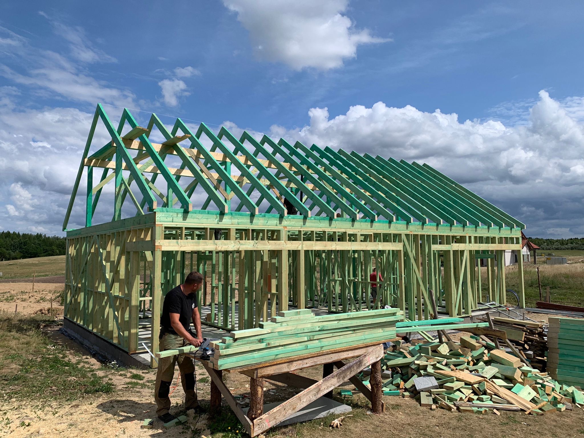 Wznoszenie szkieletu drewnianego domu jednorodzinnego: pracownik tnie deskę pilarką tarczową na tle częściowo ukończonej konstrukcji z zielonego drewna i błękitnego, pochmurnego nieba.