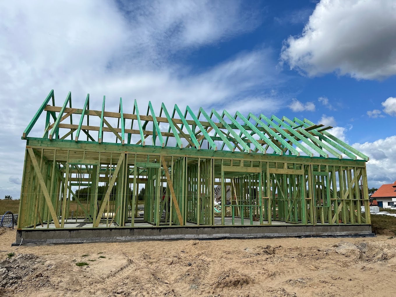 Budowa domu jednorodzinnego w stanie surowym otwartym, widoczna drewniana konstrukcja szkieletowa ścian i dachu, fundament betonowy, plac budowy z piaskiem, w tle błękitne niebo z chmurami.