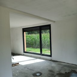 Perfect-Bud Daniel Cuch - Pokój w trakcie prac wykończeniowych, ściany zagruntowane, widoczny fragment sufitu, duże okno z widokiem na pole kukurydzy, na podłodze widoczne ślady po pracach i wiadro z masą szpachlową.