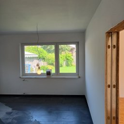 Perfect-Bud Daniel Cuch - Wnętrze pokoju w trakcie odświeżania: świeżo pomalowane białe ściany, czarna podłoga z płytek, wisząca żarówka na tle okna z widokiem na ogród, widoczna puszka farby na parapecie.