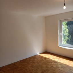 Perfect-Bud Daniel Cuch - Pusty pokój po odświeżeniu, z parkietem w jodełkę, świeżo pomalowanymi ścianami na biało i wiszącą żarówką bez klosza. Promienie słoneczne wpadają przez okno.
