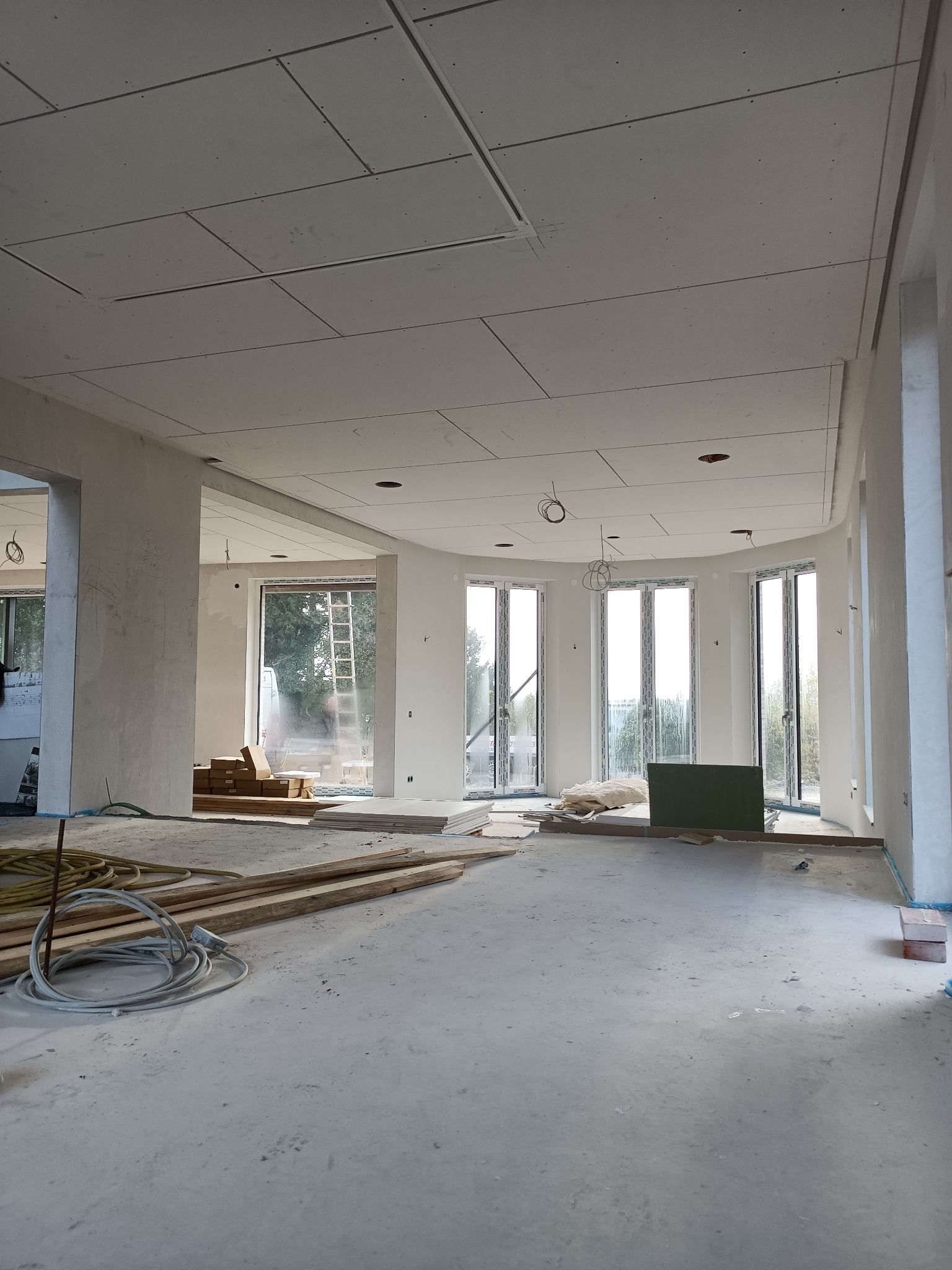Duże, jasne wnętrze w trakcie montażu sufitu z płyt kartonowo-gipsowych, widoczne przewody elektryczne, materiały budowlane i drabina w tle.