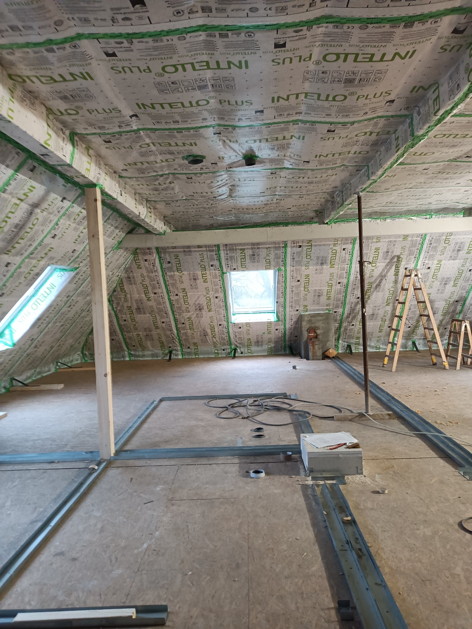 Poddasze w trakcie adaptacji, widoczne ocieplone ściany i sufit folią paroizolacyjną z zielonymi taśmami, metalowe profile pod zabudowę ścianek działowych na podłodze z płyty OSB, okno dachowe...