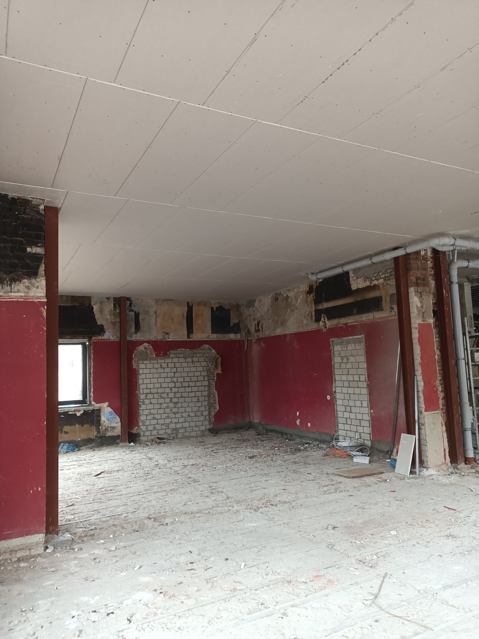 Wnętrze w trakcie prac: widoczne płyty kartonowo-gipsowe na suficie, fragmenty ceglanej ściany, czerwone ściany i elementy konstrukcyjne w surowym stanie.