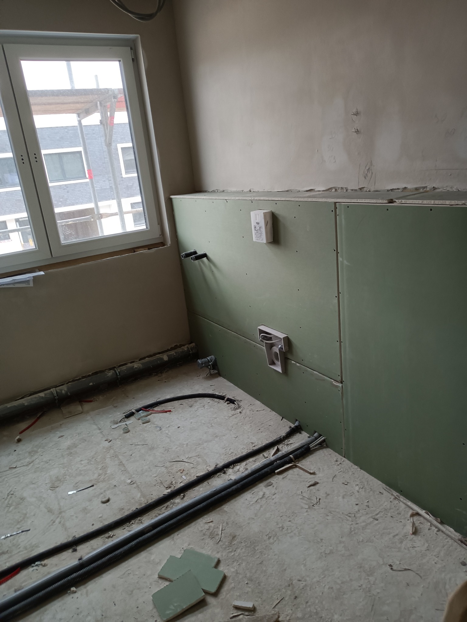 Wnętrze w trakcie prac wykończeniowych: ściana obłożona płytami kartonowo-gipsowymi, instalacja elektryczna i rury na podłodze, okno z widokiem na rusztowanie.