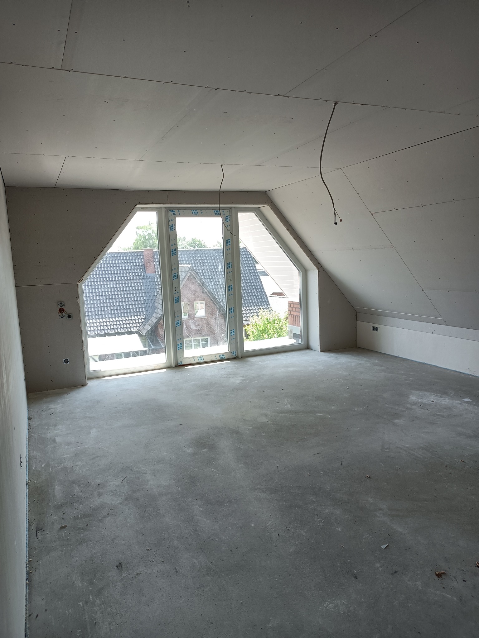 Pokój na poddaszu w trakcie remontu, ściany i sufit wyłożone płytami karton gips, widoczne okno z widokiem na domy.