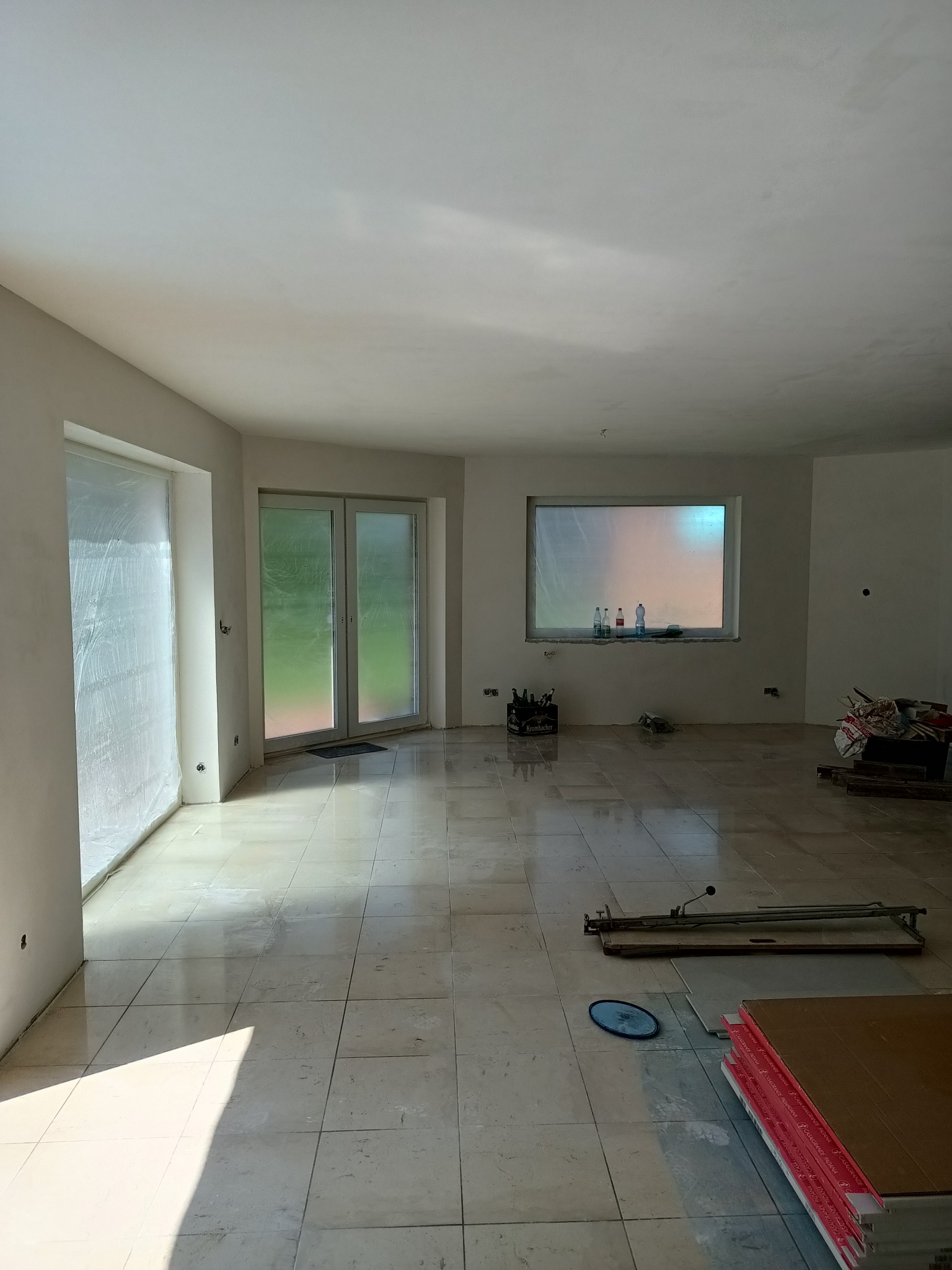 Przestronne wnętrze w trakcie prac remontowych, zabezpieczone okna folią, widoczne narzędzia do cięcia płytek i stos płyt kartonowo-gipsowych na podłodze z płytek ceramicznych.