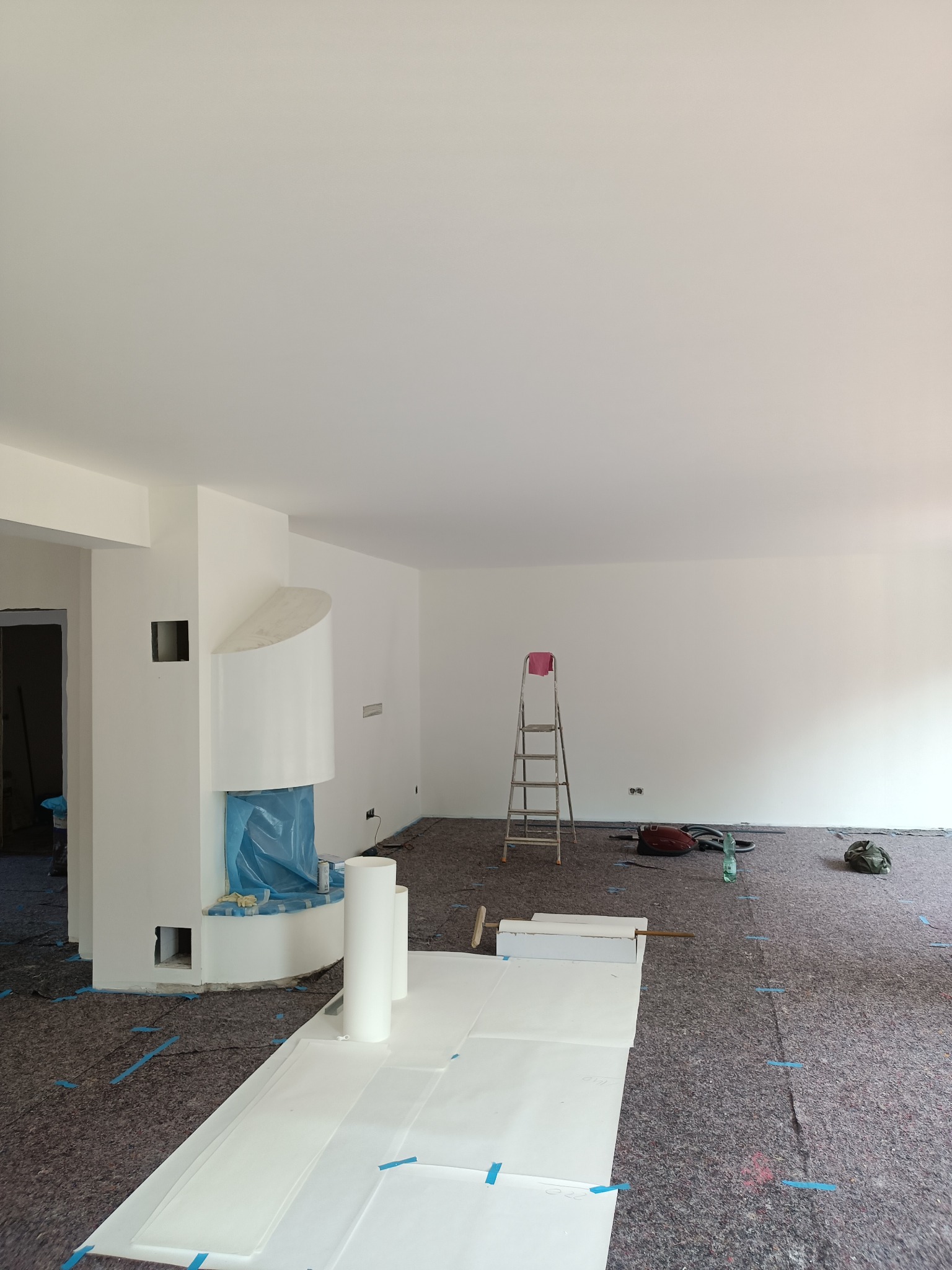 Pokój w trakcie prac przygotowawczych do malowania: zabezpieczona podłoga, osłonięty kominek folią, rozstawiona drabina, białe ściany po gładziowaniu.