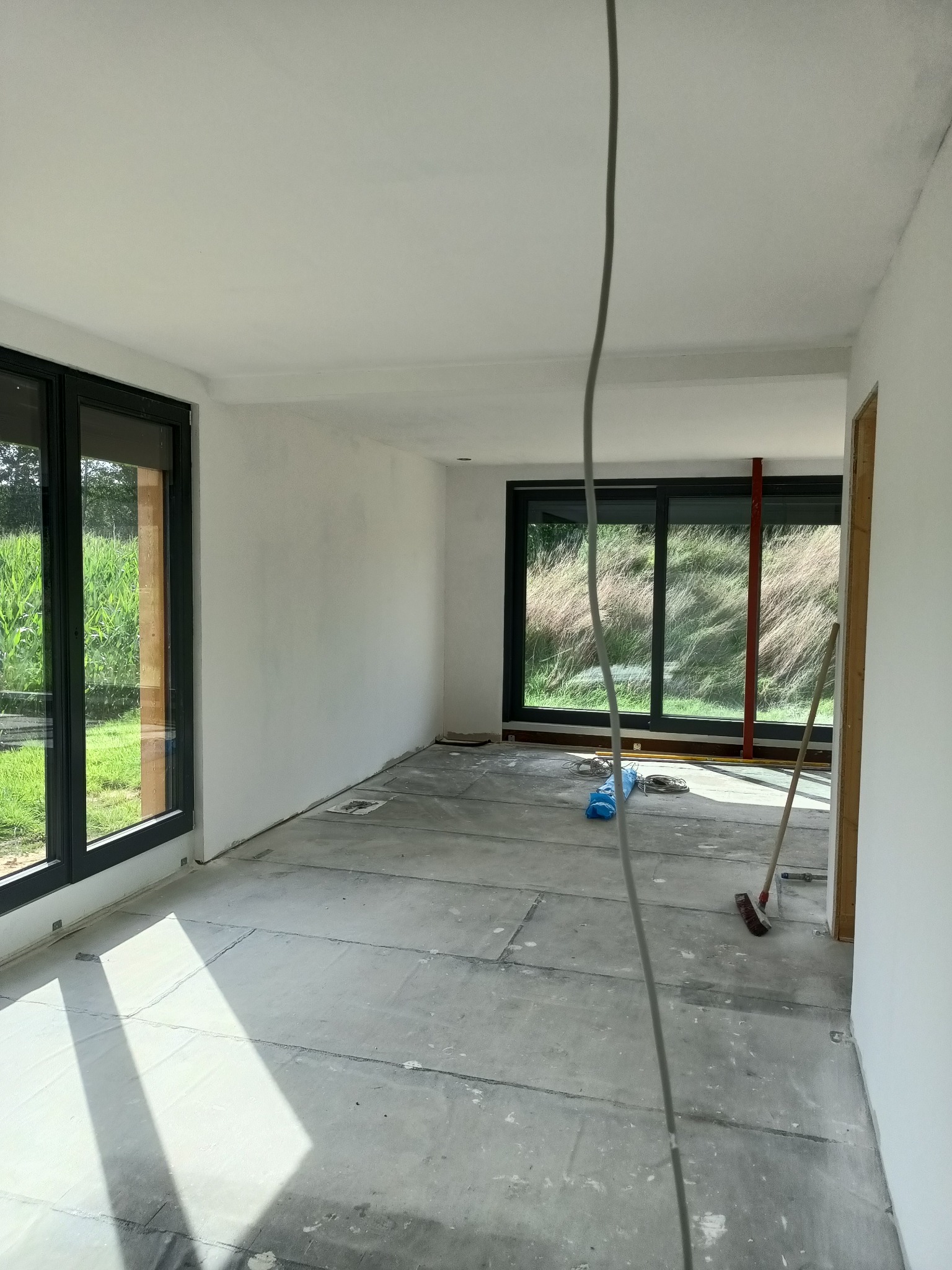 Wnętrze w trakcie prac wykończeniowych: surowe ściany, przygotowane podłoże, widoczne przewody elektryczne i narzędzia, duże okna z widokiem na zieleń.