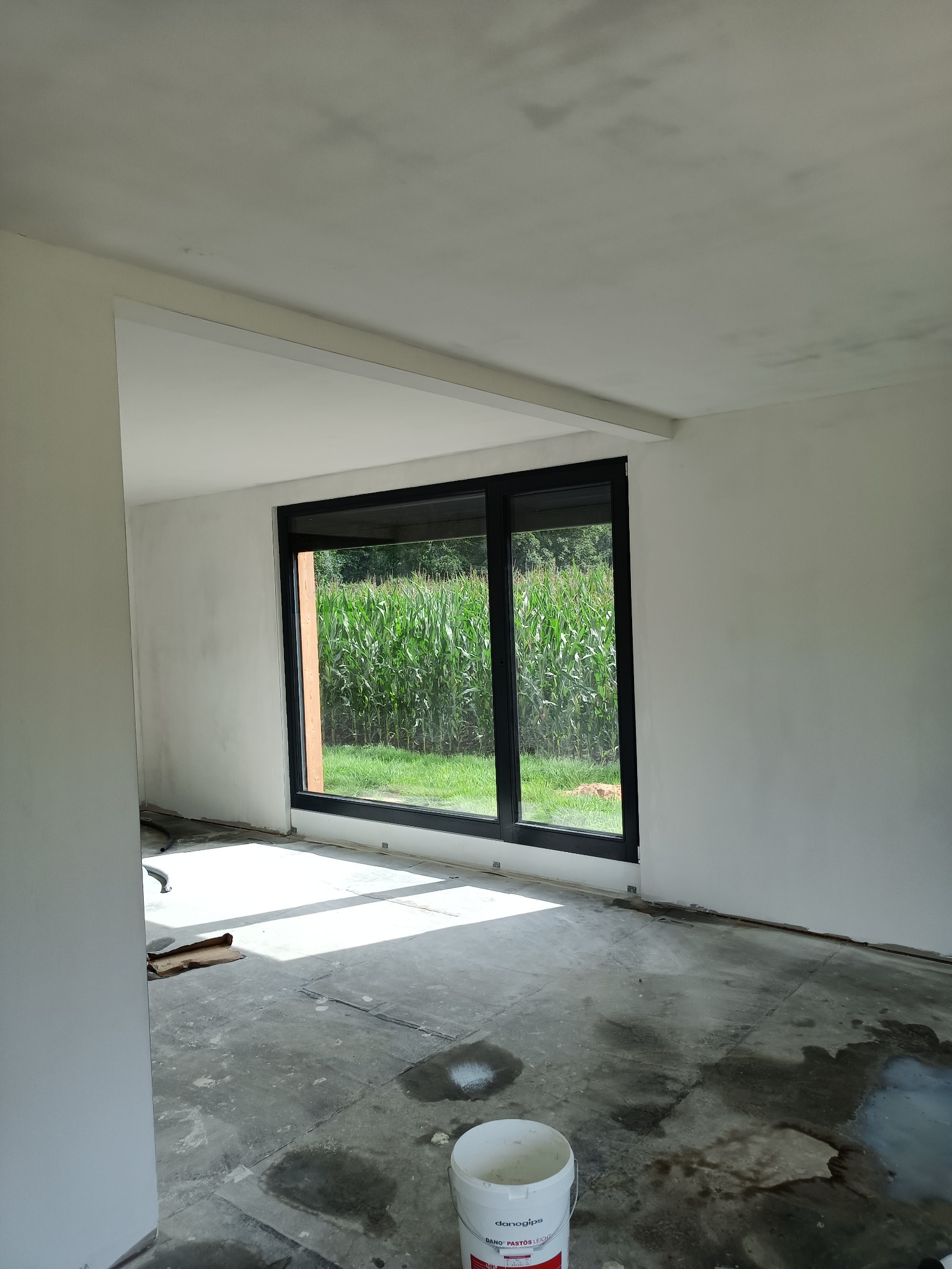 Wnętrze pokoju w trakcie prac wykończeniowych, ściany przygotowane pod malowanie, widoczne niedoskonałości na suficie, na podłodze plamy i wiadro z masą szpachlową, duże okno z widokiem na pole...