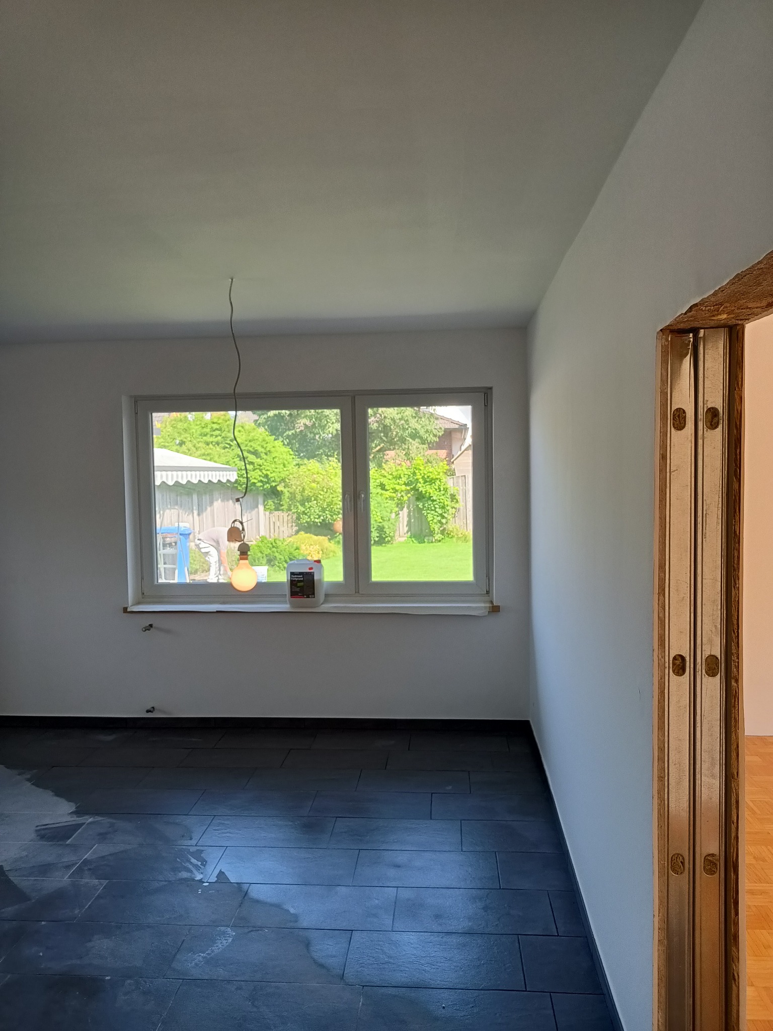 Wnętrze pokoju w trakcie odświeżania: świeżo pomalowane białe ściany, czarna podłoga z płytek, wisząca żarówka na tle okna z widokiem na ogród, widoczna puszka farby na parapecie.