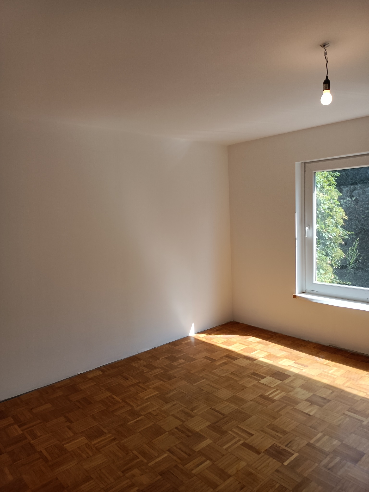 Pusty pokój po odświeżeniu, z parkietem w jodełkę, świeżo pomalowanymi ścianami na biało i wiszącą żarówką bez klosza. Promienie słoneczne wpadają przez okno.