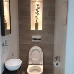 Nowoczesna łazienka z umywalką nablatową na ciemnym drewnianym blacie, toaletą z otwartą klapą i wnęką w szarej ścianie z dekoracyjną gałązką w wazonie podświetloną od góry.