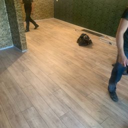 BAHMAL SP. Z.O.O - Układanie paneli podłogowych w pokoju z tapetowanymi ścianami, widoczna torba z narzędziami i fragment osoby układającej panele.