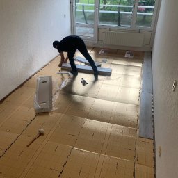 BAHMAL SP. Z.O.O - Montaż paneli podłogowych w pokoju z dużym oknem balkonowym, widoczna podkładka z nadrukiem 'easy cut', pracownik w ciemnym ubraniu przycina panel przy ścianie, obok leży gumowy młotek i karton...