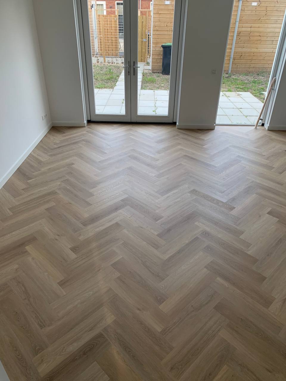 Parkiet ułożony w jodełkę francuską w jasnym, nowym wnętrzu z białymi ścianami i drzwiami balkonowymi wychodzącymi na zewnątrz.