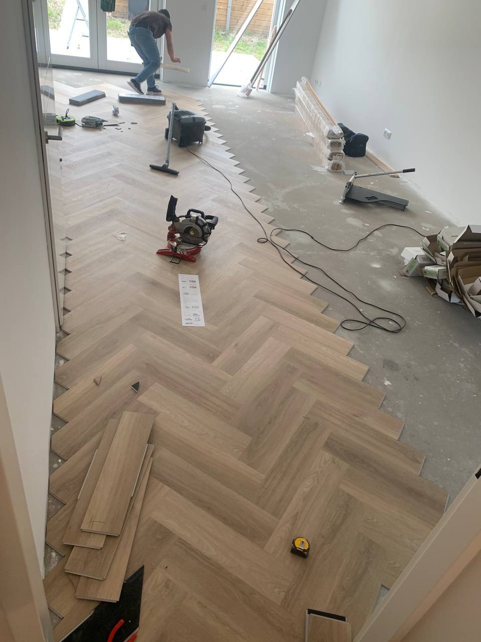 Układanie jasnych paneli podłogowych w jodełkę w trakcie remontu, widoczna piła ukośnica, odkurzacz i częściowo ułożona podłoga w surowym pomieszczeniu.