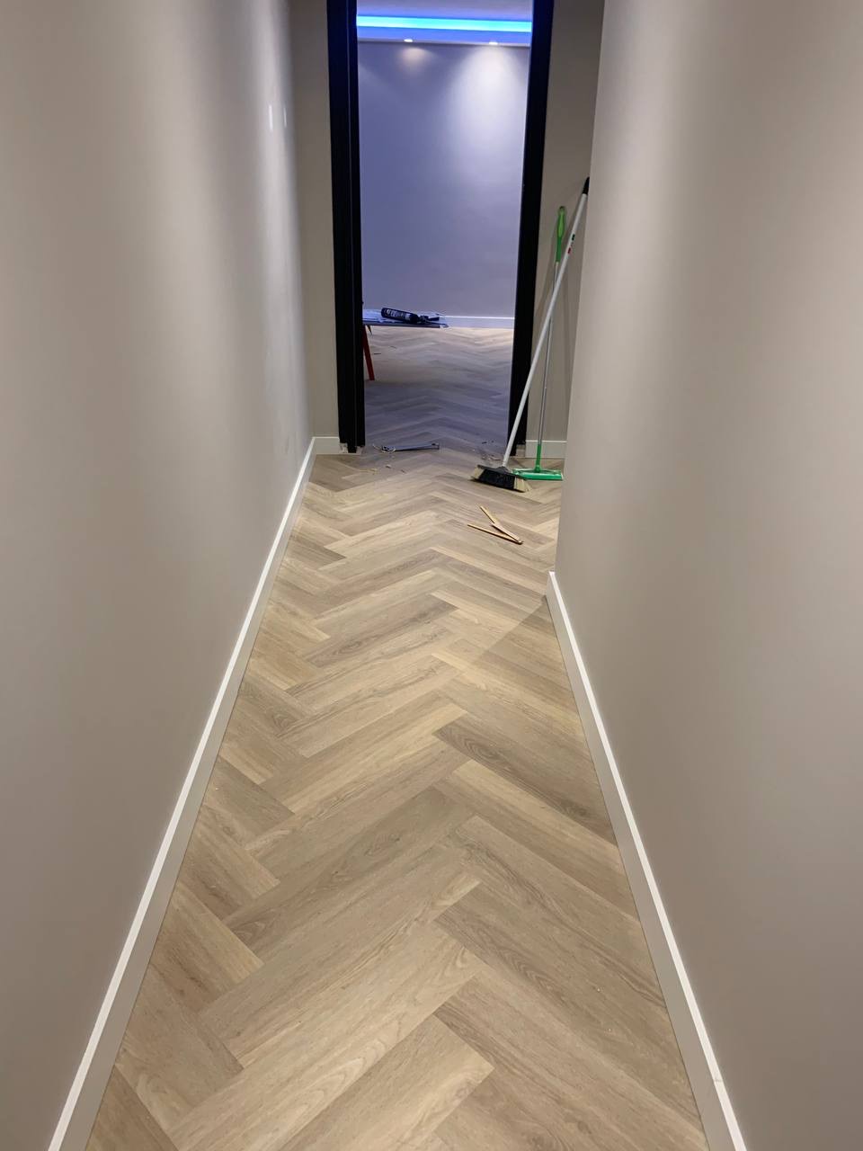 Korytarz z ułożoną parkietową podłogą w jodełkę w odcieniach beżu, widoczne listwy przypodłogowe, w tle otwarte drzwi do pokoju z niebieskim oświetleniem sufitowym, narzędzia i miotła w rogu.