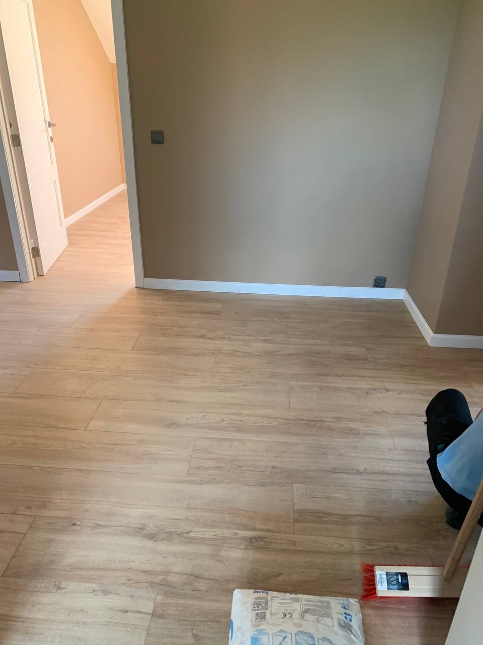 Układanie paneli podłogowych imitujących drewno w jasnym pokoju, widoczny worek kleju i szczotka do rozprowadzania kleju, częściowo widoczna osoba w roboczym ubraniu.