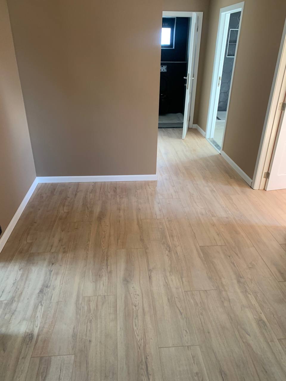 Korytarz z panelami podłogowymi imitującymi drewno, widok na otwarte drzwi do łazienki z ciemnymi płytkami i okienkiem oraz drugie drzwi do pomieszczenia z jasnymi płytkami, ściany pomalowane...