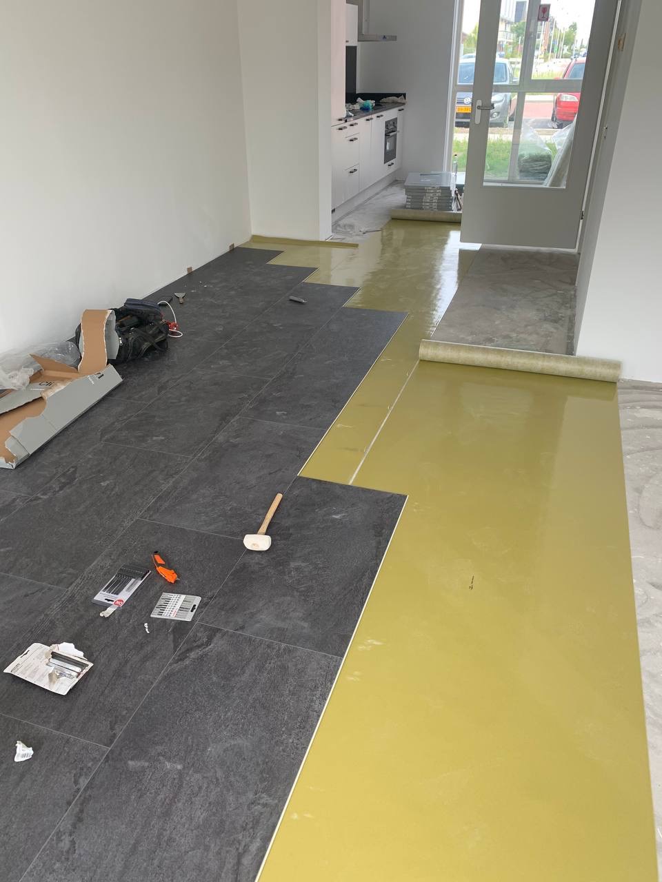 Układanie ciemnoszarych płytek na podkładzie z folii w remontowanym pomieszczeniu z widoczną kuchnią, młotek gumowy i narzędzia glazurnicze na płytkach.