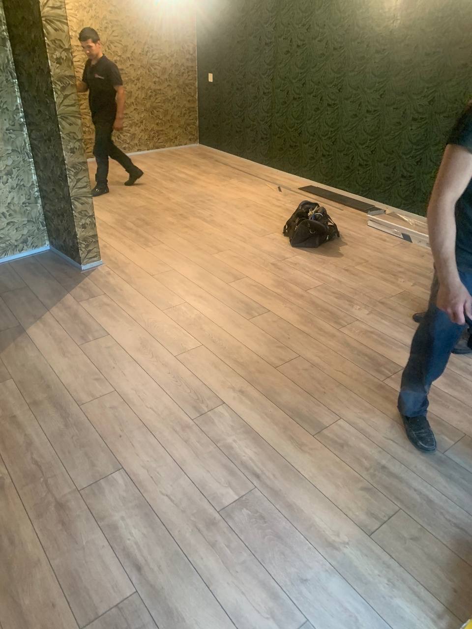 Układanie paneli podłogowych w pokoju z tapetowanymi ścianami, widoczna torba z narzędziami i fragment osoby układającej panele.