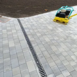 Ułożona kostka brukowa w odcieniach szarości z liniowym odwodnieniem, zagęszczarka wibrująca na powierzchni, przygotowany grunt w tle.