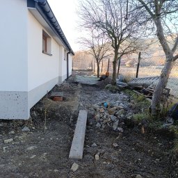 Inbruk Adam Pilch - Przygotowanie terenu pod kostkę brukową: wykop, widoczna deska, obrzeża budynku. W tle drzewa i siatka ogrodzeniowa. Poranek, lekki mróz.