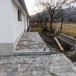 Inbruk Adam Pilch - Świeżo ułożona, szara kostka brukowa tworząca ścieżkę przy białym budynku. Widoczny młotek brukarski, ziemia i drabina w tle. Ujęcie z niskiej perspektywy.