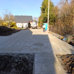 Inbruk Adam Pilch - Przygotowany podkład pod kostkę brukową na posesji. Widoczny piasek, obrzeża, ziemia i narzędzia. W tle dom i pracownik. Słoneczny dzień.