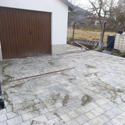 Inbruk Adam Pilch - Szara kostka brukowa ułożona przed garażem z brązową bramą. Widoczna kratka odpływowa i resztki piasku na kostce. W tle drabina i zbiorniki na wodę.