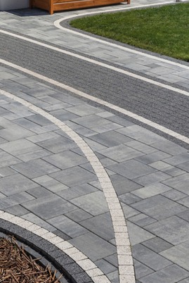 Nowoczesna nawierzchnia z kostki brukowej w odcieniach szarości, z dekoracyjnymi łukami z jaśniejszej kostki i pasem z drobnej kostki, obrzeże z ciemnej kostki, trawnik w tle.