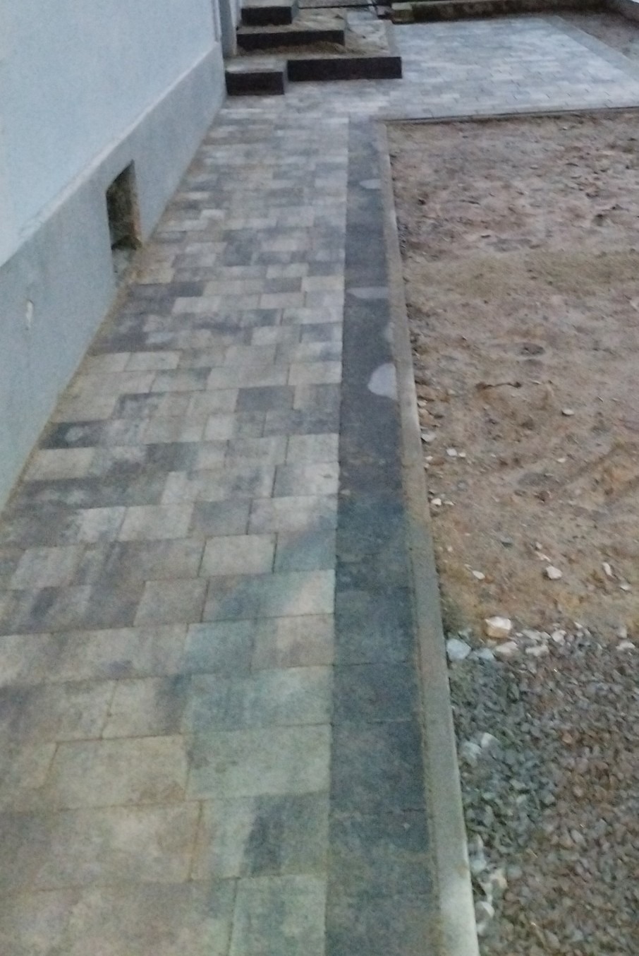 Ścieżka z szarej kostki brukowej prowadząca wzdłuż ściany budynku, obok fragmentu terenu przygotowanego pod dalsze prace brukarskie.