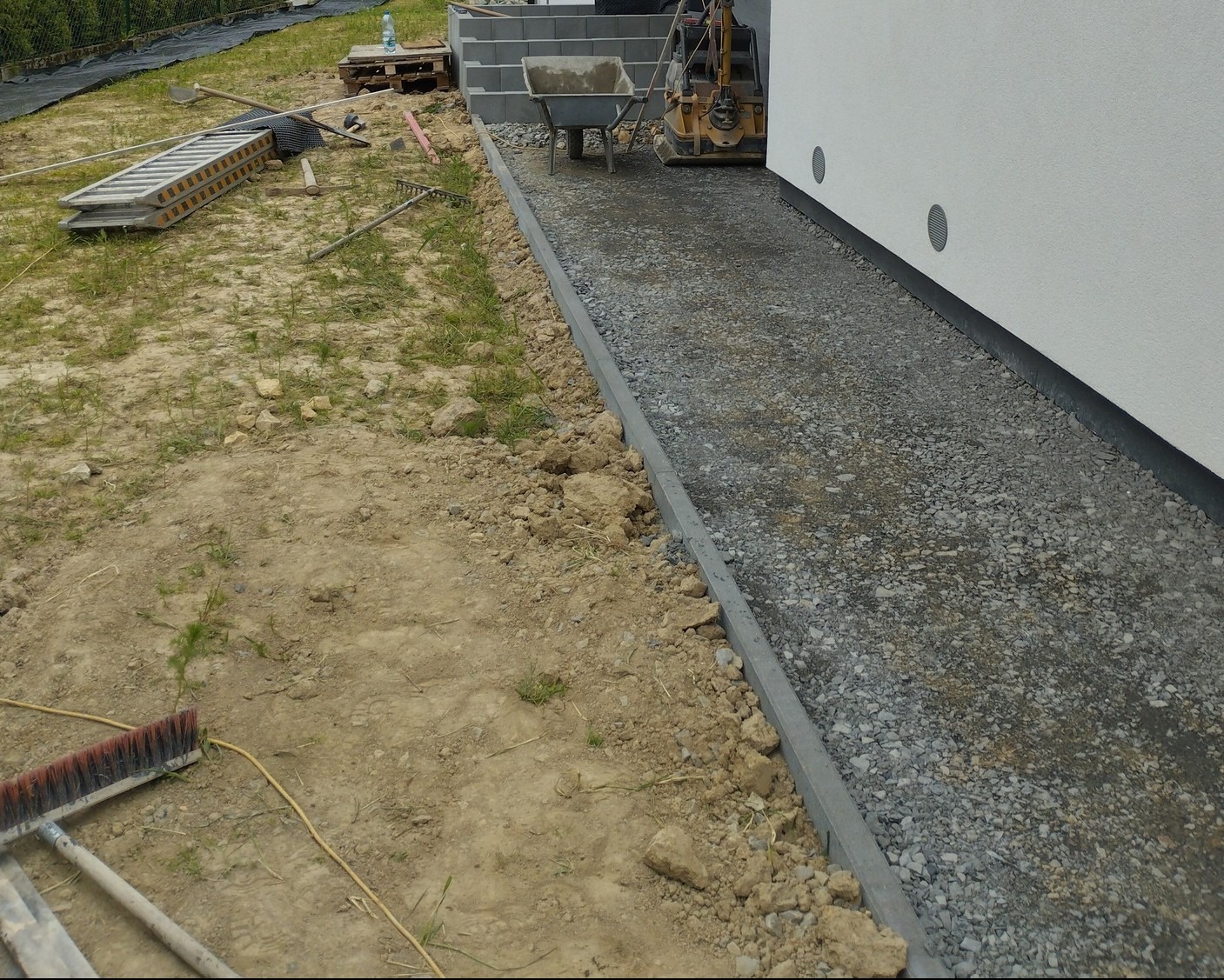 Przygotowanie podłoża pod kostkę brukową z widocznymi narzędziami i ubijarką w pobliżu budynku.