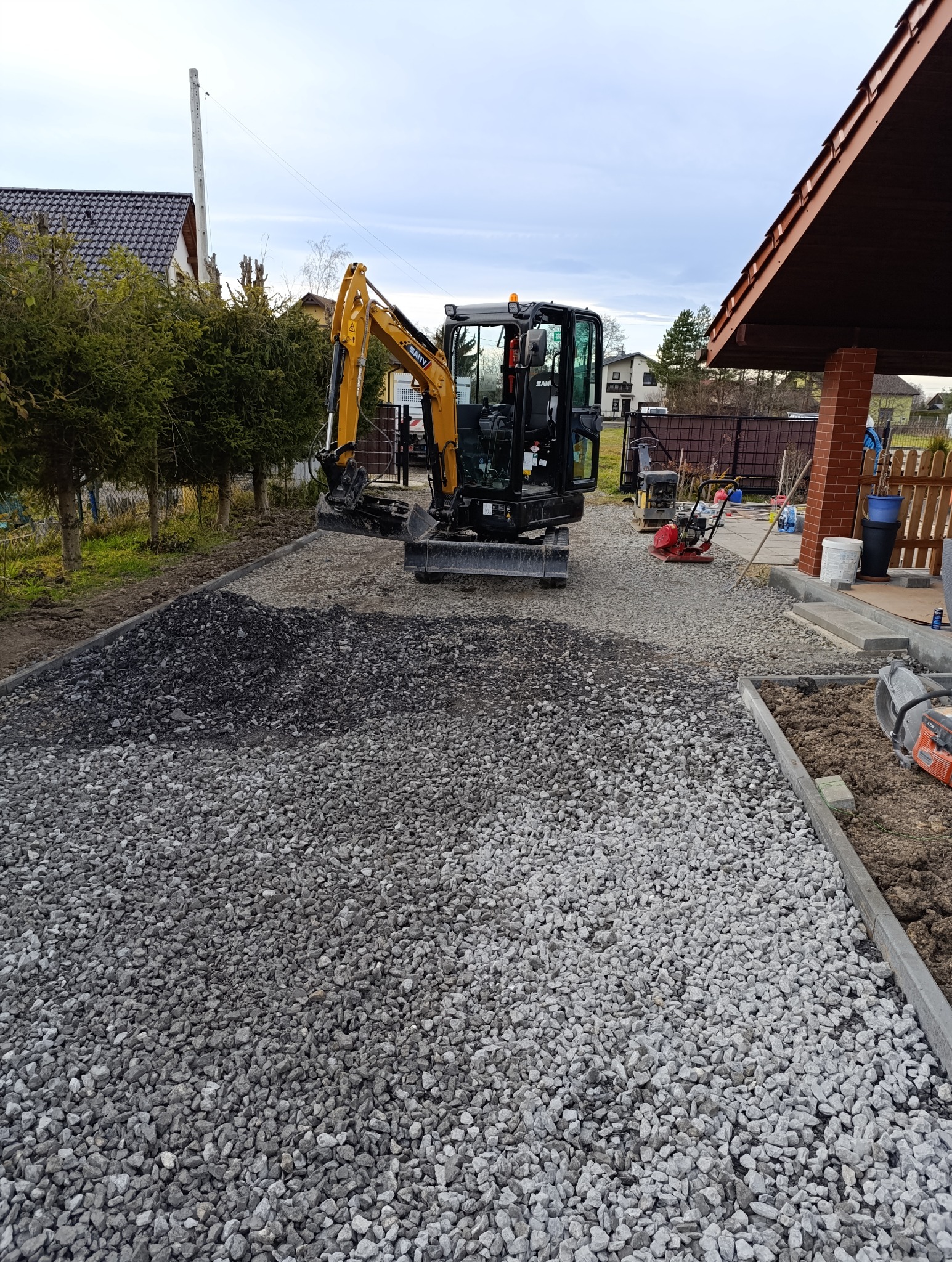 Mini koparka SANY na podsypce z kruszywa, przygotowanie podłoża pod nawierzchnię, w tle zagęszczarki i dom jednorodzinny.