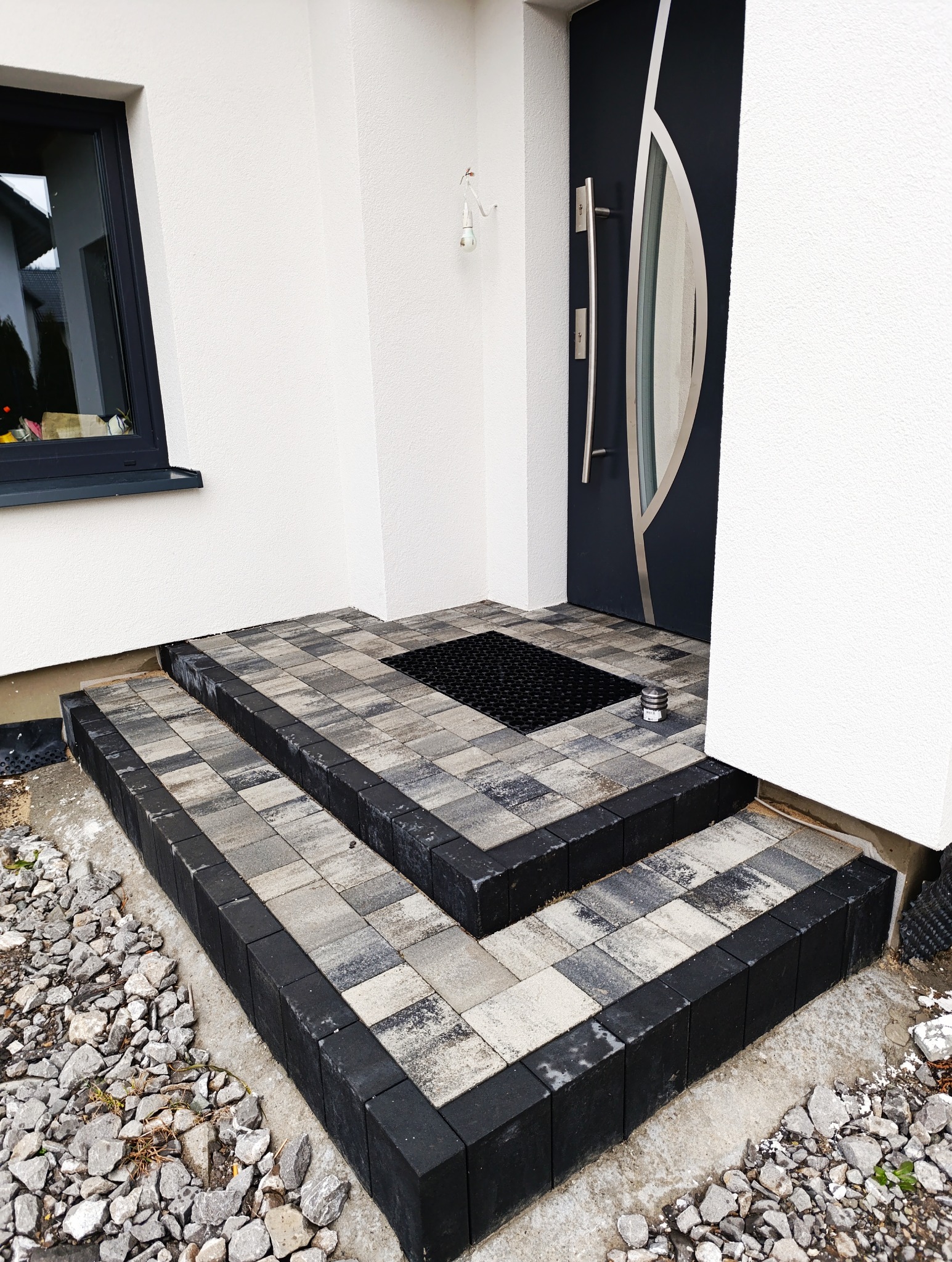 Schody wejściowe do domu wyłożone kostką brukową w odcieniach szarości z czarnym obramowaniem, widoczny fragment elewacji z oknem i czarne drzwi wejściowe z elementami stali nierdzewnej.