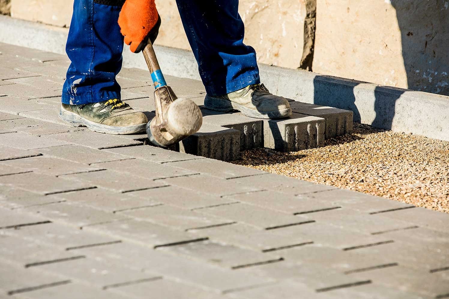 Pracownik w pomarańczowych rękawicach dobija gumowym młotkiem szarą kostkę brukową na podsypce z drobnego żwiru, widoczne buty robocze i fragment nieotynkowanego muru.