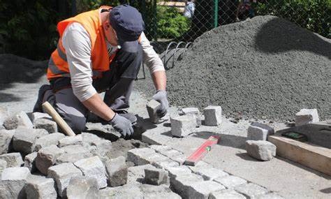 Pracownik w pomarańczowej kamizelce układa kostkę granitową na podsypce z piasku; w tle hałda kruszywa.