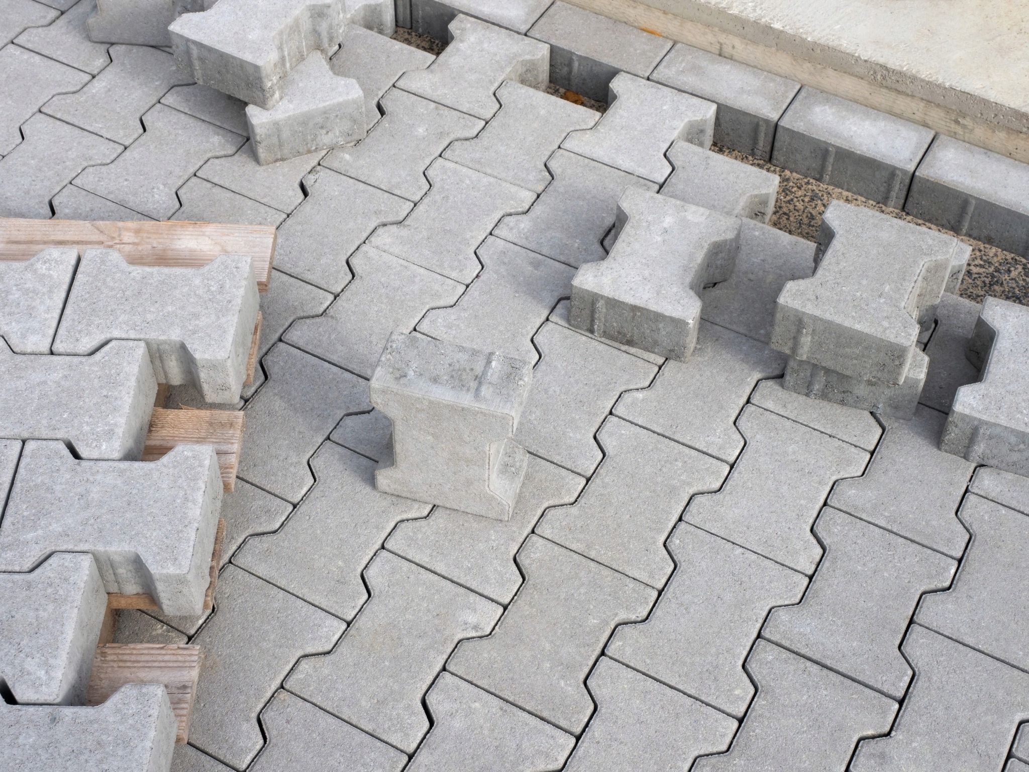 Szare, betonowe kostki w kształcie litery H układane na piasku, widoczne przerwy między niektórymi elementami, paleta z kostką obok