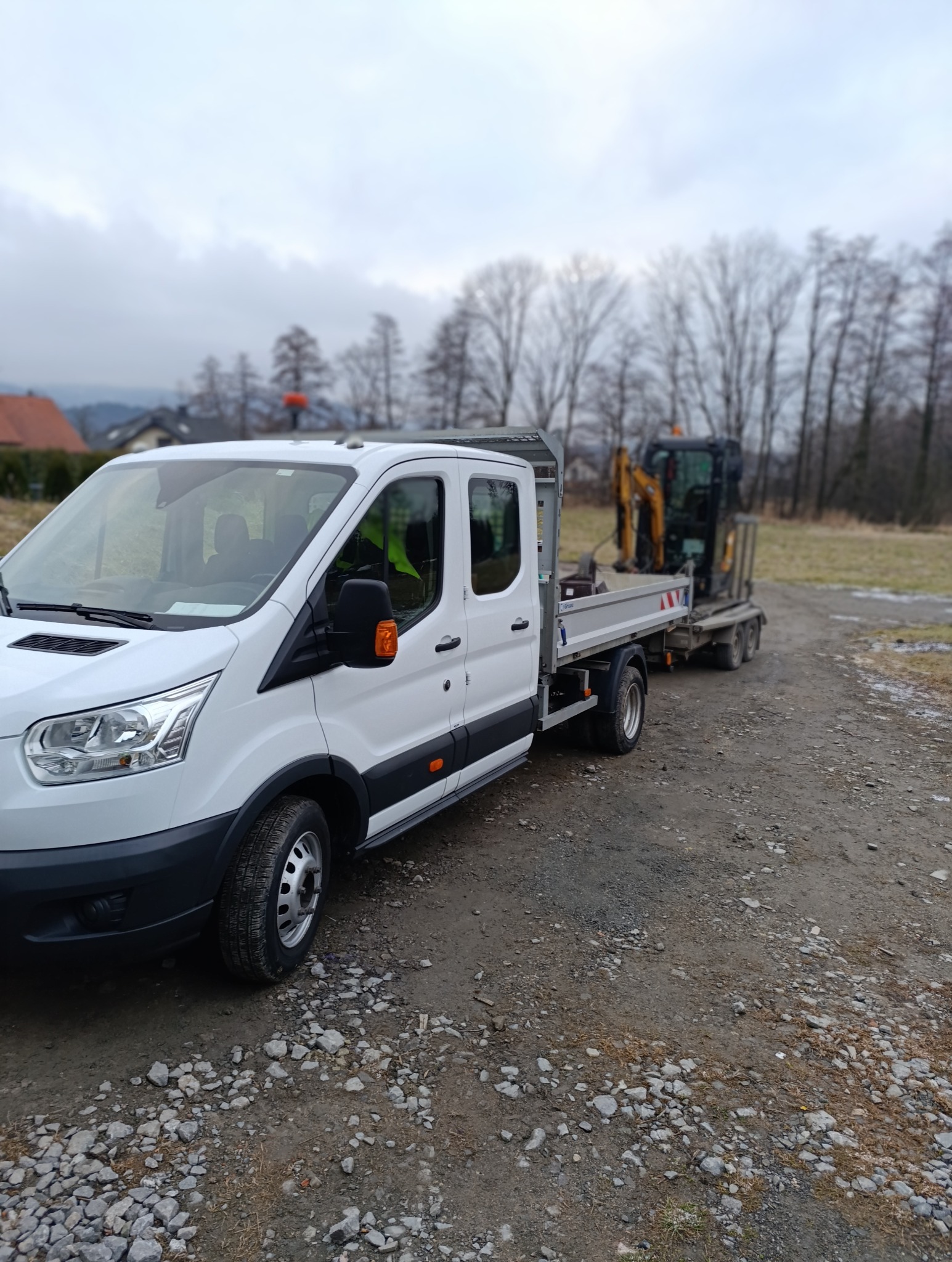 Biały samochód dostawczy z platformą transportową ciągnie małą koparkę na tle szarego nieba i drzew bez liści.