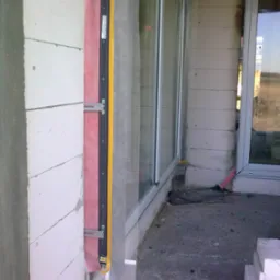 Fragment ściany z widocznym ociepleniem wokół okna w trakcie montażu, z zastosowaniem różowej wełny mineralnej i żółtej listwy uszczelniającej.