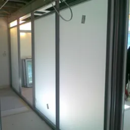 Wnętrze biurowe z zamontowanymi ściankami działowymi z aluminium i mlecznego szkła, widoczne kable pod sufitem i surowa podłoga w trakcie wykończenia.