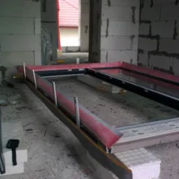 Przygotowanie ramy okiennej do montażu w surowym murze, widoczne elementy uszczelniające i podkłady izolacyjne, w tle fragment dachu budynku.