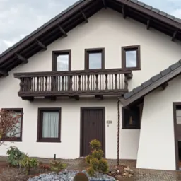 Dwukondygnacyjny dom z jasną elewacją, ciemnobrązowymi oknami, balkonem z balustradą i numerem domu 149, otoczony zielenią i kamieniami.