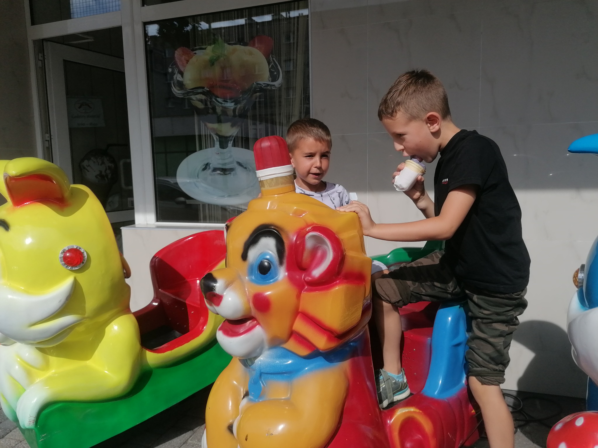 Dwaj chłopcy bawią się na kolorowych, plastikowych automatach do gier przed lodziarnią, jeden z nich je lody z papierowego kubka.
