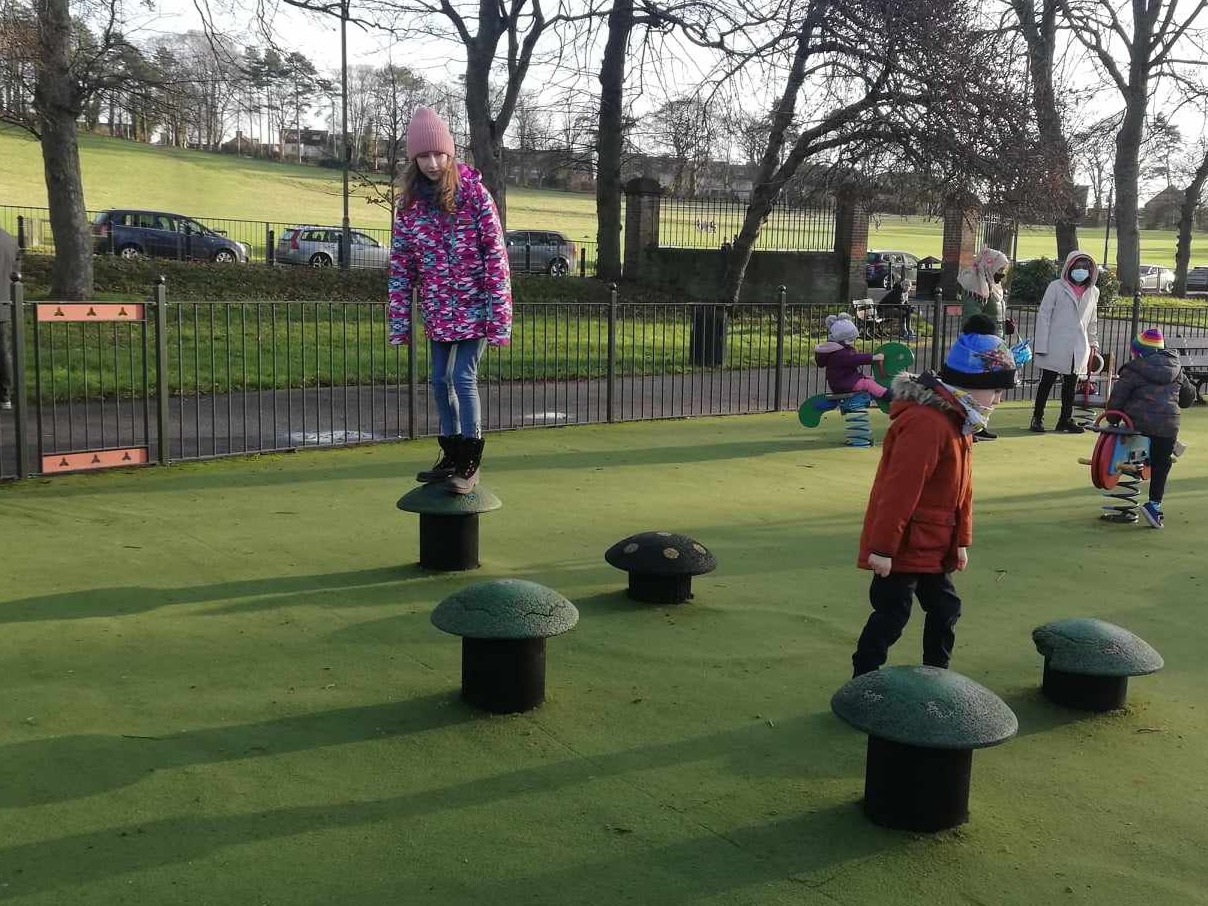 Dziewczynka w różowej czapce i kurtce stoi na grzybku na placu zabaw w Łukawcu, w tle widoczni inni bawiący się ludzie.