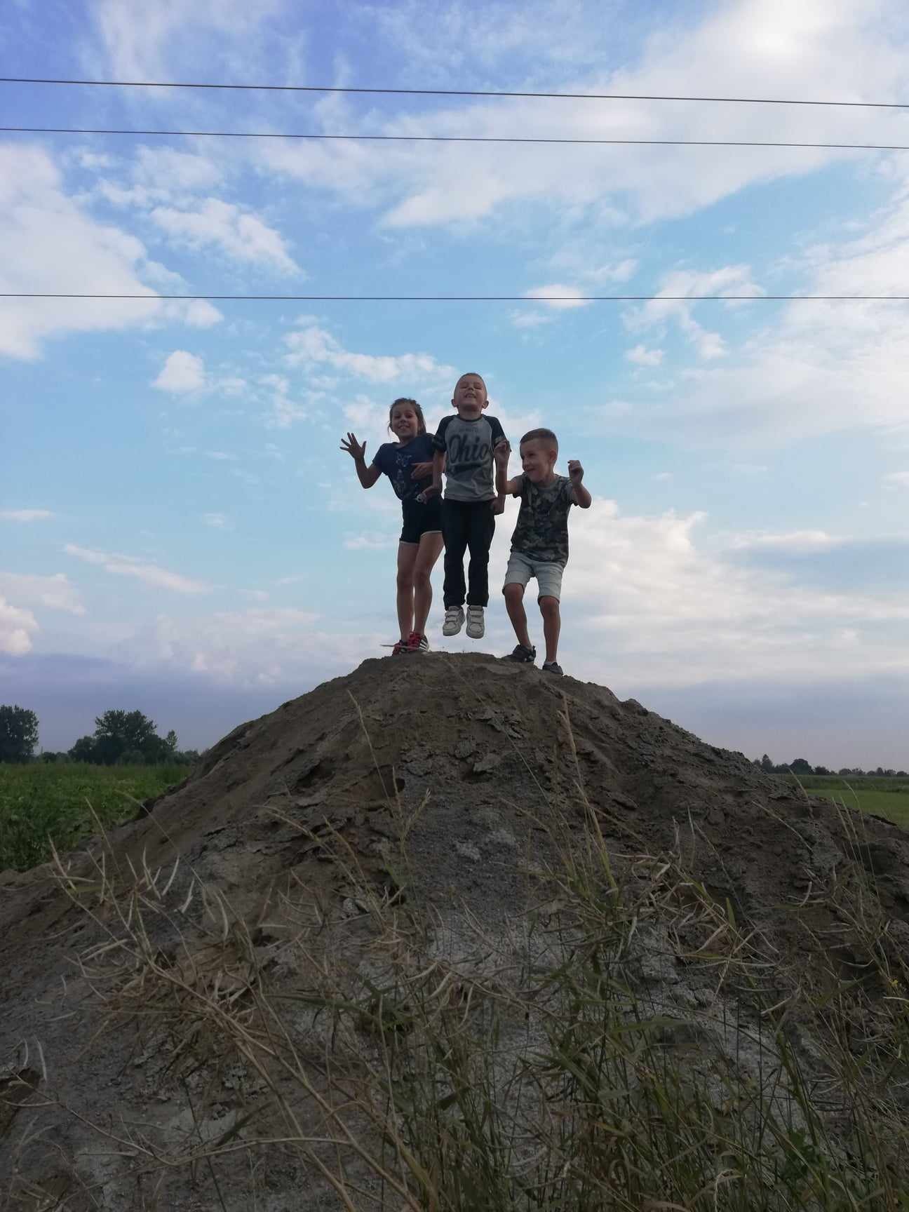 Troje roześmianych dzieci skacze z pryzmy ziemi na tle błękitnego nieba z białymi obłokami i polami uprawnymi w oddali.