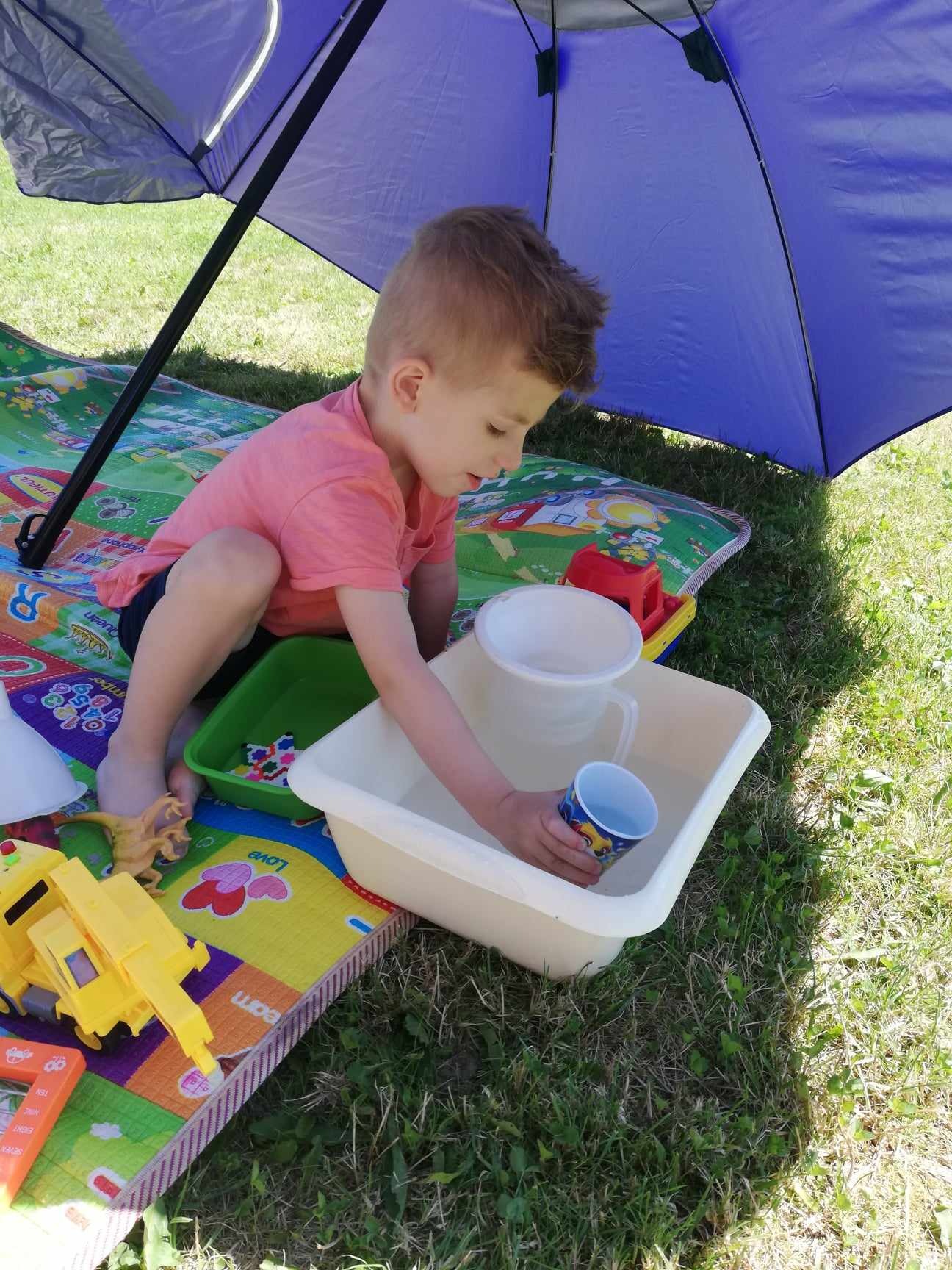 Chłopiec bawi się wodą w wanience pod niebieskim parasolem na trawie, otoczony zabawkami na kolorowej macie edukacyjnej.